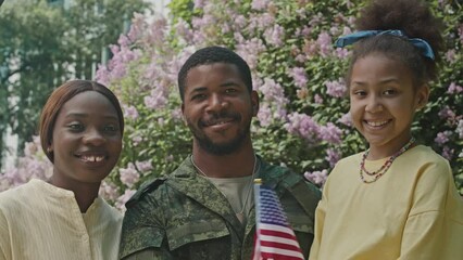 Portrait of happy African American soldier in military uniform holding US flag and posing for camera with joyous wife and little daughter outdoors beside tree in blossom