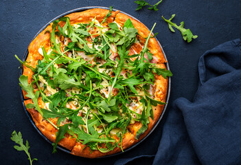 Hot pizza with mozzarella cheese, ham, pesto and fresh arugula, black table background, top view