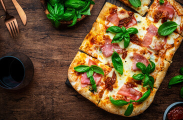 Hot Roman pizza with ham, pancetta, mozzarella cheese, sun dried tomatoes, sauce and green basil, rustic wooden table background, top view