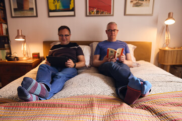 Same sex older male couple reading and relaxing on bed in apartment