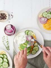 Hands with knife and fork at the table cutting omelet on a plate with avocado, cucumber salad and red onion. Yogurt sauce seasoned with thyme, lemon and black pepper.