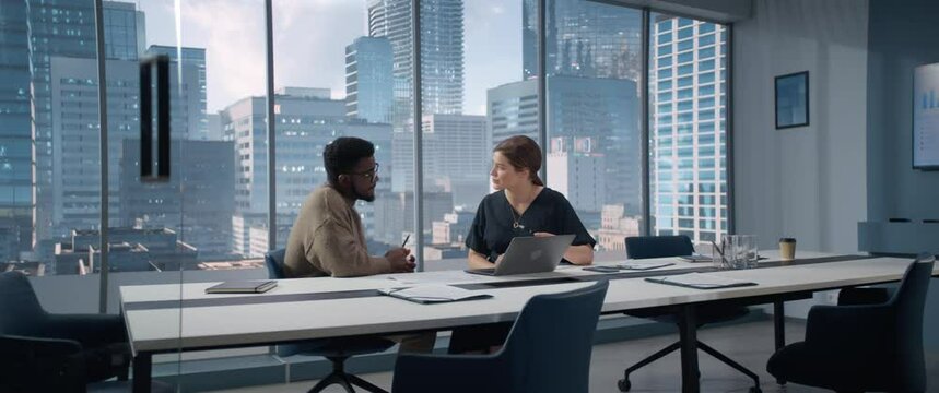 Two Multiethnic Colleagues Have a Discussion in Bright Modern Skyscraper Office in an Agency. Balck Creative Director and Female Project Manager Talking About Data On Laptop Computer. Anamorphic Shot