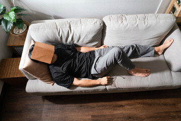 Man with mental health problem lying on comfortable couch and covering face with book to sleeping