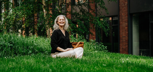Portrait of beautiful smiling woman with gray hair sitting on the grass in city park.