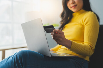 Young Asian woman holding credit card and smartphone sitting on sofa at home doing online banking transactions in home online shopping in living room. online payment and ordering concept.