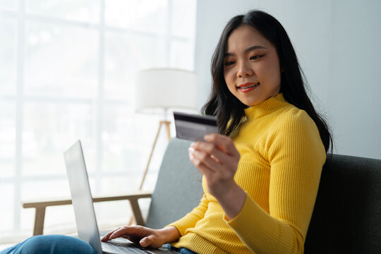 Young Asian woman holding credit card and smartphone sitting on sofa at home doing online banking transactions in home online shopping in living room. online payment and ordering concept.