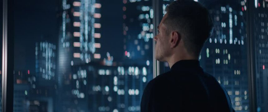 Close Up Portrait of Caucasian Businessman in a Suit Standing Next to Window in Big City Office with Skyscrapers Late At Night. Male General Manager Looking Out Of Window Thoughtfully. Anamorphic Shot