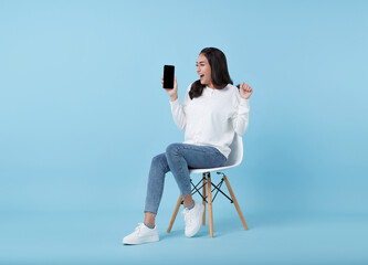 Happy young Asian woman showing empty screen smartphone. While her sitting on white chair isolated on blue studio background.