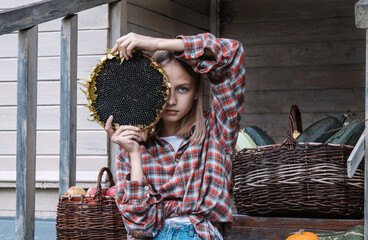 Girl is sitting on steps of an old village house holding large sunflower near her face. Ripe pumpkins lie nearby and there are baskets filled with harvested crops: apples and zucchini. Fall, autumn