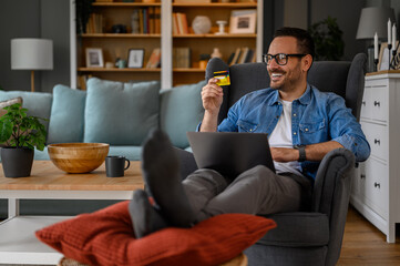 Happy man doing online shopping on laptop paying with credit card while sitting in home office