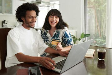 Cute brunette woman with credit card and her boyfriend looking through goods on laptop screen and choosing something in online shop