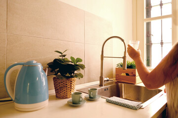 bright kitchen, woman gathering clean water into glass for drinking from modern chrome faucet,...