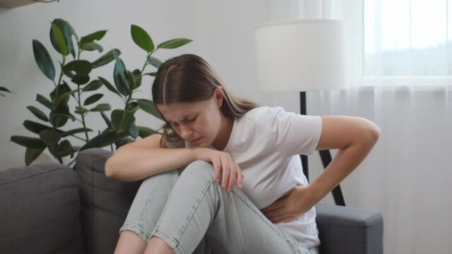 Unhealthy young caucasian woman suffering from stomach ache sit on sofa. Upset brunette girl holding belly and feeling abdominal menstrual pain or bowel and digestion problems. Food poisoning concept