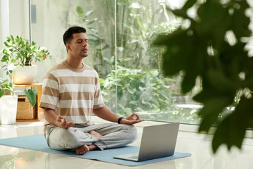 Foto op Plexiglas Young serene man in activewear keeping eyes closed during home yoga practice while sitting on mat in pose of lotus in front of laptop screen © DragonImages