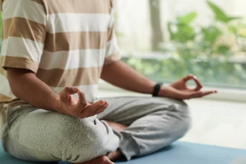 Poster Close-up of young active man in t-shirt and sweatpants sitting on the floor in pose of lotus while practicing yoga in home environment © DragonImages
