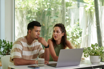 Obraz premium Young intercultural couple sitting by desk in front of laptop and discussing working point while serious man in striped t-shirt looking at screen
