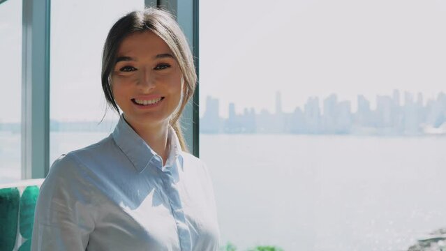 Portrait of happy successful young Caucasian woman working at modern company and standing near window in beautiful office. Attractive charming elegant businesswoman in suit looking at camera.