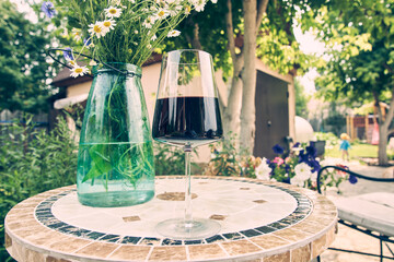 In the backyard of the cottage there is a table with a glass of wine and a bouquet of daisies in a glass vase. The concept of a country holiday on the weekend. High quality photo