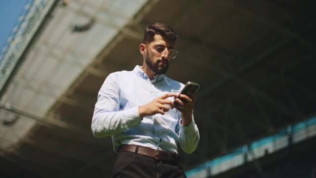 Businessman in formal wear standing on sport stadium, looking on mobile phone and winning money. Concept of sport, fan, betting and finances, gambling, bookmaker