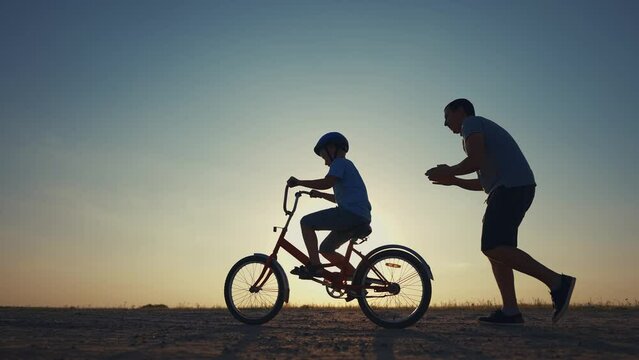family play in the park. father teaching son to ride a bike. happy family kid dream concept. son learn to ride a bike silhouette. sunset father supporting child son riding bike summer in the park