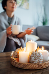 Focus on autumn fall cozy mood decor composition with a blurred woman covered plaid selecting movie with remote controller and drinking tea on the sofa. Movie night party at home. Cozy relaxing time.