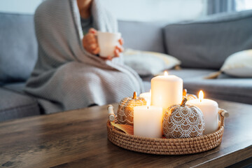 Focus on autumn fall cozy mood decor composition. Orange pumpkins, burning candles with a blurred woman covered with plaid and drinking tea on the sofa. Movie night party at home. Cozy relaxing time.