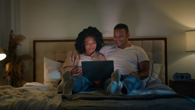 African American couple family man and woman in bed night home dark bedroom evening watching laptop tv movie watch film relaxing romantic date love relationship computer video shopping online together