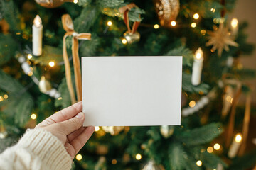 Hand holding empty greeting card on background of stylish decorated christmas tree with golden lights. Christmas card mock up. Space for text. Season greetings template © sonyachny
