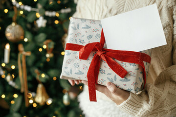 Hands holding empty greeting card and stylish wrapped christmas gift against festive christmas tree with lights. Christmas card mock up. Space for text. Season greetings postcard template © sonyachny