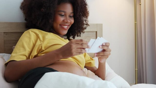 Black African American pregnant woman looking her ultrasound scan photos on bed. Expecting mother waiting her baby. Concept of pregnancy, Maternity prenatal care.