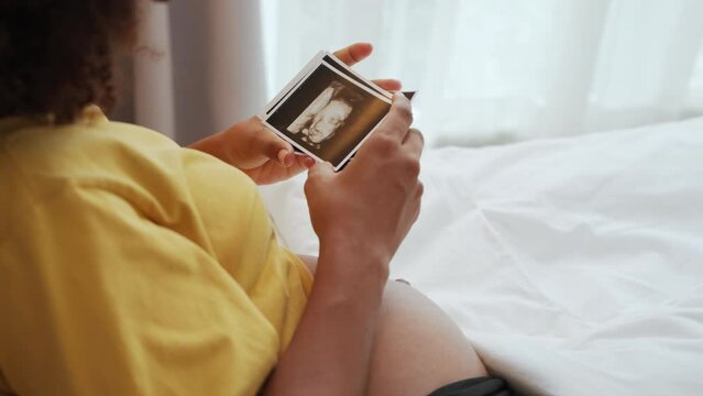 Black African American pregnant woman looking her ultrasound scan photos on bed. Expecting mother waiting her baby. Concept of pregnancy, Maternity prenatal care.