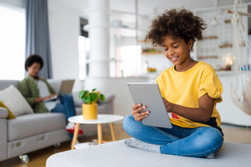 Carefree smiling African American young girl with curly hair using digital tablet sitting with...