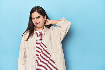 Young Caucasian woman on blue backdrop touching back of head, thinking and making a choice.