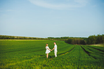 Naklejka na ściany i meble Pregnant couple hugging in a green spring field. Expectant parents in the park of white flowering trees. A romantic couple expecting a baby. A walk through a green field