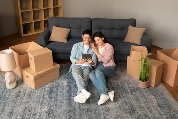 Fototapeta na wymiar Asian Couple Using Digital Tablet Sitting Among Moving Boxes Indoor