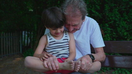 Loving family moment of grandfather bonding with grandson seated at park bench during summer day....