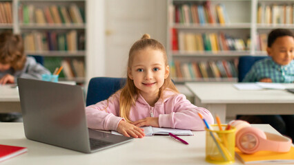 Happy caucasian school girl using laptop at class, looking and smiling at camera, using technology during tech lesson