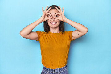 Young caucasian woman on blue backdrop showing okay sign over eyes