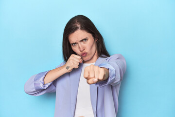 Woman in blue blazer on blue background throwing a punch, anger, fighting due to an argument, boxing.