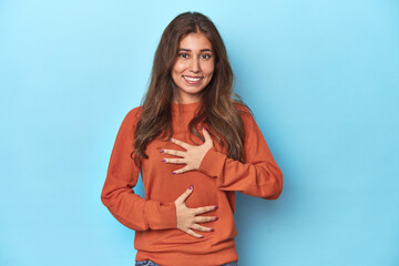 Teen girl in vibrant orange sweater on blue laughs happily and has fun keeping hands on stomach.