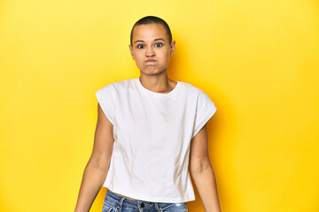 Shaved head woman in white tank top, yellow backdrop blows cheeks, has tired expression. Facial...