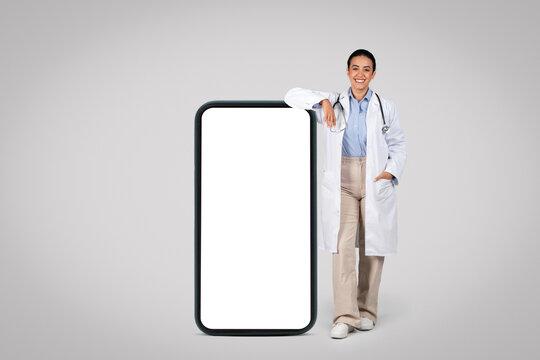 Cheerful latin female doctor leaning on huge smartphone with blank screen, grey background, mockup, full length