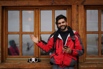 Handsome dark haired guy waving with his hand while holding mobile phone. Male wearing red ski suit...