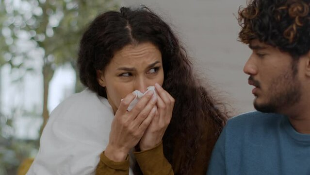 Young upset sick middle eastern woman blowing her nose and sneezing at home, her caring husband giving her pills