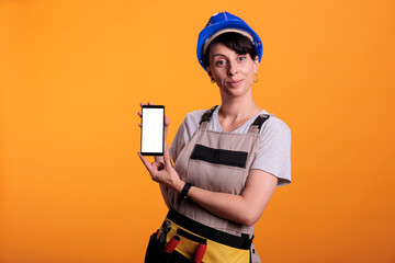 Female contractor showing blank white screen on mobile phone display, advertising isolated mockup template. Happy construction worker using copyspace on smartphone, studio shot.