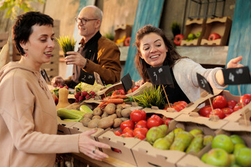 Positive market stand owner showing organic produce to woman, presenting colorful bio fruits and vegetables. Young farmer selling homegrown eco products from personal local farm.