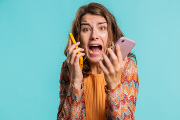 Irritated nervous stressed young woman talking screaming on two mobile phones having conversation conflict quarrel complaint dispute discuss solve problem. Girl isolated on blue studio background
