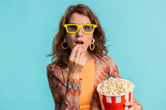 Excited young woman in 3D glasses eating popcorn and watching interesting tv serial, sport game, film, online social media movie content. Brunette lovely girl isolated on blue studio background