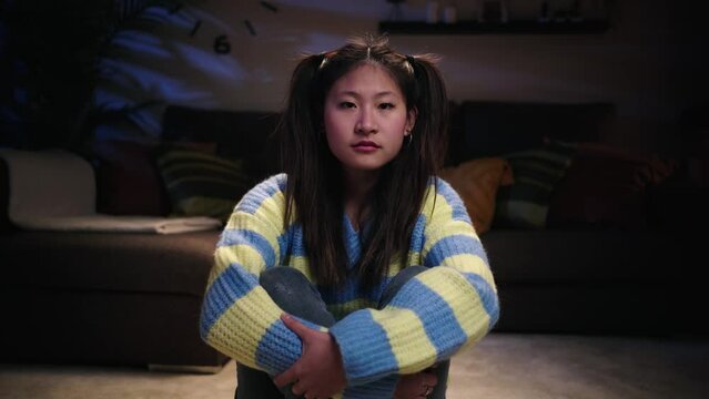 Portrait of serious young Asian woman sitting in living room at home. Chinese girl looking earnest at camera. Sad female indoors at night with concerned aptitude. Generation z people and copy space