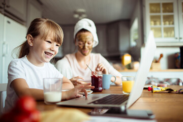 Young mother and daughter having breakfast and using a laptop in the morning in the kitchen